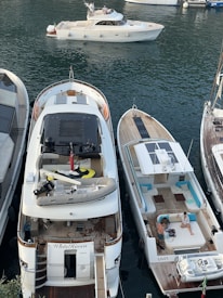 Several boats are docked at a marina. A luxurious yacht named 'WhiteHaven' features an upper deck with a dinghy and other equipment. Next to it, a smaller boat has people lounging and socializing on its deck. The water appears calm and there is another boat sailing through the marina in the background.