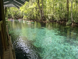 A serene river setting with crystal-clear water reflecting sunlight, surrounded by lush green trees. Several people are leisurely floating on colorful inflatables in the water. The wooden structure of a dock or building is visible in the foreground.
