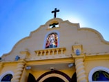 A church façade with a prominent cross at the top. The building has a beige exterior with decorative elements. There is a stained glass window featuring a religious figure positioned in the center above the entrance. The sky in the background is clear and blue.