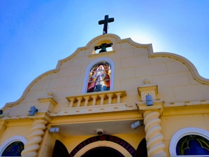 A church façade with a prominent cross at the top. The building has a beige exterior with decorative elements. There is a stained glass window featuring a religious figure positioned in the center above the entrance. The sky in the background is clear and blue.