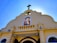 A church façade with a prominent cross at the top. The building has a beige exterior with decorative elements. There is a stained glass window featuring a religious figure positioned in the center above the entrance. The sky in the background is clear and blue.