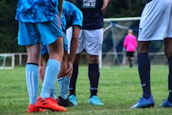 A group of soccer players are on a grassy field. One player is bent over, adjusting their shin guard while others stand around. The players are wearing uniforms in shades of blue, and a goalkeeper in a bright pink jersey is visible near the goal in the background.