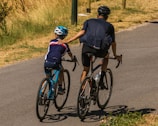 A man and a child are cycling on a paved road surrounded by dry grass. The man gently holds the child's shoulder as they ride side by side. Both are wearing helmets and sporty cycling outfits.