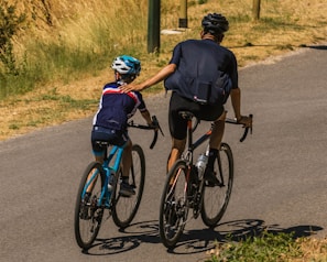 A man and a child are cycling on a paved road surrounded by dry grass. The man gently holds the child's shoulder as they ride side by side. Both are wearing helmets and sporty cycling outfits.