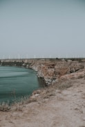 A series of wind turbines line the horizon above steep, rocky cliffs overlooking a body of water. The landscape is natural and expansive, with sparse vegetation on the cliff edges and a clear, pale sky overhead.
