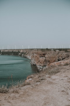 A series of wind turbines line the horizon above steep, rocky cliffs overlooking a body of water. The landscape is natural and expansive, with sparse vegetation on the cliff edges and a clear, pale sky overhead.