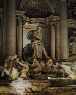A detailed stone sculpture featuring a large central figure with a flowing beard and draped clothing, surrounded by other smaller figures and mythical creatures. The backdrop includes tall columns and architectural elements with intricate carvings and inscriptions, all in a warm, illuminated setting which emphasizes the textures of the stone.