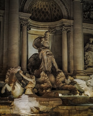 A detailed stone sculpture featuring a large central figure with a flowing beard and draped clothing, surrounded by other smaller figures and mythical creatures. The backdrop includes tall columns and architectural elements with intricate carvings and inscriptions, all in a warm, illuminated setting which emphasizes the textures of the stone.