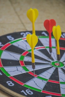 A dartboard with several darts embedded in it is depicted, with three yellow darts and one red dart. The dartboard's circular sections are clearly visible, showing different colored segments including black, white, red, and green.