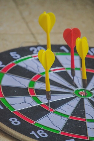 A dartboard with several darts embedded in it is depicted, with three yellow darts and one red dart. The dartboard's circular sections are clearly visible, showing different colored segments including black, white, red, and green.