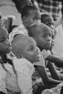 A group of children sitting closely together, engaged and attentive. They appear to be in a classroom or similar environment, focused on something off-camera.