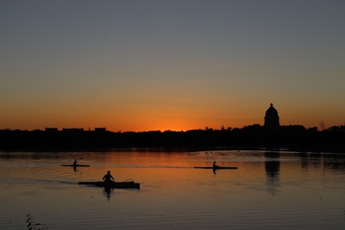 Silhouetted against a vibrant sunset, several kayakers paddle across a calm body of water. In the distance, the outline of a large dome-shaped building is visible on the horizon, surrounded by trees and low buildings. The water reflects the warm hues of the sunset, adding depth to the scene.