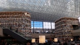 A large and modern library with tall, curved bookshelves filled with numerous books. There is a central area with tables and large lamps, and an escalator on the left side. The ceiling is made up of glass panels, allowing natural light to illuminate the space. There are people scattered throughout the library, reading and walking.
