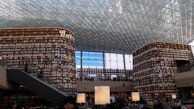 A large and modern library with tall, curved bookshelves filled with numerous books. There is a central area with tables and large lamps, and an escalator on the left side. The ceiling is made up of glass panels, allowing natural light to illuminate the space. There are people scattered throughout the library, reading and walking.