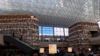 A large and modern library with tall, curved bookshelves filled with numerous books. There is a central area with tables and large lamps, and an escalator on the left side. The ceiling is made up of glass panels, allowing natural light to illuminate the space. There are people scattered throughout the library, reading and walking.