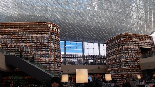 A large and modern library with tall, curved bookshelves filled with numerous books. There is a central area with tables and large lamps, and an escalator on the left side. The ceiling is made up of glass panels, allowing natural light to illuminate the space. There are people scattered throughout the library, reading and walking.
