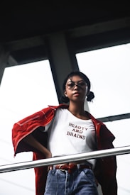 A person stands confidently in front of a window, wearing glasses and a white shirt with the words 'BEAUTY AND BRAINS' printed on it. Over the shirt is a red jacket draped over the shoulders, and the person is wearing blue jeans with a red belt while holding onto a railing in a modern, industrial setting.
