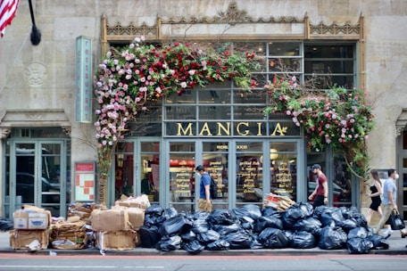 An urban street scene in front of a restaurant named Mangia, which features an elaborate floral decoration with pink and red flowers around the entrance. In front of the restaurant, there is a large pile of black garbage bags and cardboard boxes. Several people are walking past the entrance on the sidewalk.