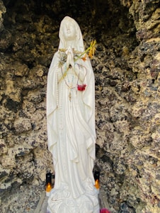 A white statue of a figure in a robe stands with hands joined in prayer, adorned with multiple rosaries. Flowers and candles are placed around its base, and the statue is set against a rugged stone background.