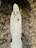 A white statue of a figure in a robe stands with hands joined in prayer, adorned with multiple rosaries. Flowers and candles are placed around its base, and the statue is set against a rugged stone background.