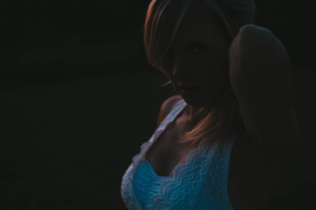 A woman with blonde hair is wearing a white lace top. The image is dark, with low lighting, creating a mysterious and shadowy effect. The woman's face is partially obscured by shadows.