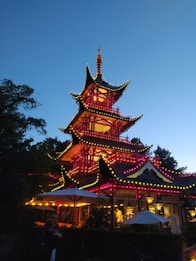 A traditional multi-tiered pagoda is illuminated with vibrant string lights against a clear evening sky. The structure is adorned with red and yellow lights, creating a festive and lively atmosphere. Surrounding the pagoda are outdoor umbrellas and garden foliage, with people appearing to enjoy the ambiance.