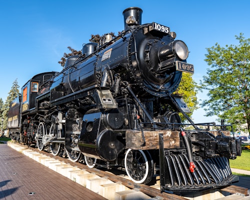A black steam locomotive is prominently displayed on a section of track in an outdoor setting. The locomotive has shiny, well-maintained metal surfaces with white-rimmed wheels and several visible mechanical components. Behind the locomotive, lush green trees can be seen against a bright blue sky, indicating a clear day.