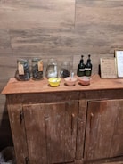 A rustic wooden cabinet is topped with several glass jars filled with herbs or leaves, two pumps perhaps containing liquid soap or lotion, and three small bowls with spoons inside them. A framed sign is also displayed, and the backdrop features wooden plank walls.