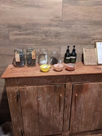 A rustic wooden cabinet is topped with several glass jars filled with herbs or leaves, two pumps perhaps containing liquid soap or lotion, and three small bowls with spoons inside them. A framed sign is also displayed, and the backdrop features wooden plank walls.