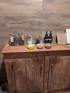 A rustic wooden cabinet is topped with several glass jars filled with herbs or leaves, two pumps perhaps containing liquid soap or lotion, and three small bowls with spoons inside them. A framed sign is also displayed, and the backdrop features wooden plank walls.