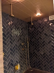 A modern shower area with dark blue herringbone-patterned tiles covering the walls. It features two ceiling-mounted rainfall showerheads, a handheld shower, gold-colored fixtures, and a wall-mounted shelf holding three bottles, possibly containing soap or shampoo. The ceiling has warm recessed lighting and an air vent.