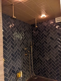 A modern shower area with dark blue herringbone-patterned tiles covering the walls. It features two ceiling-mounted rainfall showerheads, a handheld shower, gold-colored fixtures, and a wall-mounted shelf holding three bottles, possibly containing soap or shampoo. The ceiling has warm recessed lighting and an air vent.