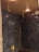 A modern shower area with dark blue herringbone-patterned tiles covering the walls. It features two ceiling-mounted rainfall showerheads, a handheld shower, gold-colored fixtures, and a wall-mounted shelf holding three bottles, possibly containing soap or shampoo. The ceiling has warm recessed lighting and an air vent.