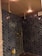 A modern shower area with dark blue herringbone-patterned tiles covering the walls. It features two ceiling-mounted rainfall showerheads, a handheld shower, gold-colored fixtures, and a wall-mounted shelf holding three bottles, possibly containing soap or shampoo. The ceiling has warm recessed lighting and an air vent.