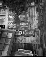 A collection of old, weathered wooden signs with faded lettering is mounted on a rustic wooden wall. The signs advertise estate sales, liquidations, and antiques. Several vintage window frames lean against the wall, adding to the eclectic and nostalgic feel. The scene includes patches of foliage partially covering some of the signs.