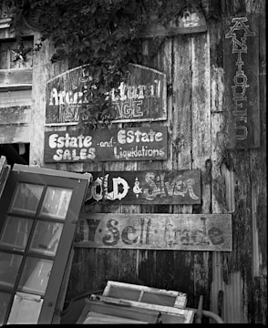 A collection of old, weathered wooden signs with faded lettering is mounted on a rustic wooden wall. The signs advertise estate sales, liquidations, and antiques. Several vintage window frames lean against the wall, adding to the eclectic and nostalgic feel. The scene includes patches of foliage partially covering some of the signs.