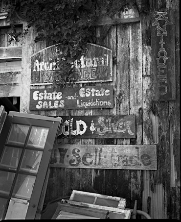 A collection of old, weathered wooden signs with faded lettering is mounted on a rustic wooden wall. The signs advertise estate sales, liquidations, and antiques. Several vintage window frames lean against the wall, adding to the eclectic and nostalgic feel. The scene includes patches of foliage partially covering some of the signs.