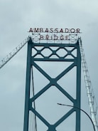 A section of a large suspension bridge with a metal lattice structure and the words 'AMBASSADOR BRIDGE' displayed at the top in bold, red lettering.