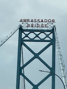 A section of a large suspension bridge with a metal lattice structure and the words 'AMBASSADOR BRIDGE' displayed at the top in bold, red lettering.