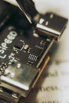 Close-up view of a microcontroller board with visible electronic components and connectors. Features include a clearly detailed circuit board with multiple pins and a partially visible brand logo. The background contains blurred text, placing focus on the intricate design of the board.