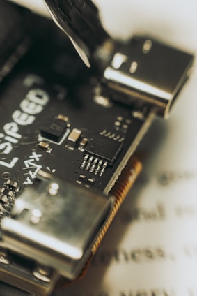 Close-up view of a microcontroller board with visible electronic components and connectors. Features include a clearly detailed circuit board with multiple pins and a partially visible brand logo. The background contains blurred text, placing focus on the intricate design of the board.