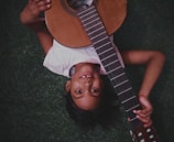 A young child lies on their back on green grass, holding a classical guitar. The child is smiling, with an expression of happiness and playfulness, and their short dark hair fans out around their head. The guitar is positioned diagonally across their torso, showcasing its strings and wooden body.