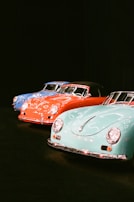 A lineup of classic cars is displayed in a dimly lit setting. Three vintage vehicles, each with smooth, rounded designs and chrome accents, are positioned side by side. The cars are painted in vibrant colors: blue, red, and aqua. Their polished surfaces reflect the ambient light, emphasizing their glossy finishes.