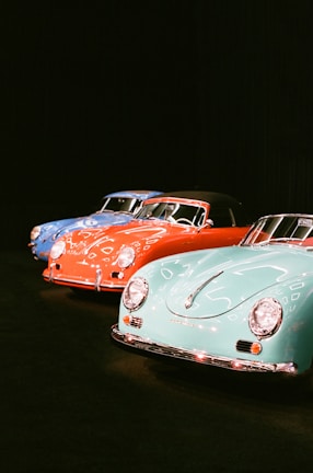 A lineup of classic cars is displayed in a dimly lit setting. Three vintage vehicles, each with smooth, rounded designs and chrome accents, are positioned side by side. The cars are painted in vibrant colors: blue, red, and aqua. Their polished surfaces reflect the ambient light, emphasizing their glossy finishes.