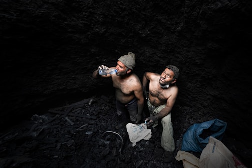 Two shirtless men covered in soot are sitting against the dark earthen walls of a mine. The men appear exhausted and are drinking water from plastic bottles. Debris and bags lie around them.