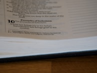 A page from a book is open, showing text from the Proverbs of Solomon. It includes a verse about the joy and grief brought by a wise or foolish son to his parents. The page is partially blurred and the book is lying on a wooden surface.