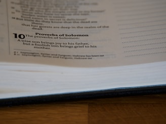 A page from a book is open, showing text from the Proverbs of Solomon. It includes a verse about the joy and grief brought by a wise or foolish son to his parents. The page is partially blurred and the book is lying on a wooden surface.