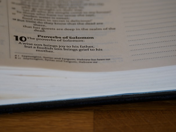 A page from a book is open, showing text from the Proverbs of Solomon. It includes a verse about the joy and grief brought by a wise or foolish son to his parents. The page is partially blurred and the book is lying on a wooden surface.