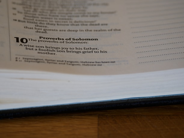 A page from a book is open, showing text from the Proverbs of Solomon. It includes a verse about the joy and grief brought by a wise or foolish son to his parents. The page is partially blurred and the book is lying on a wooden surface.