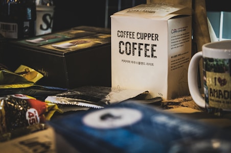 A cluttered table filled with various coffee-related items, featuring a box labeled 'Coffee Cupper Coffee' in the center. There are other packages and a mug with text and an image. The setting appears to be a cozy, indoor environment.